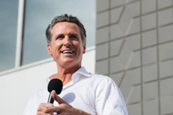 Man in white shirt speaking at microphone with California flag in background