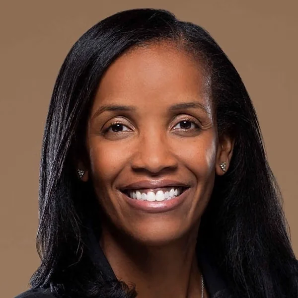 Professional headshot of a smiling woman with long black hair wearing small earrings against a tan background