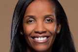 Professional headshot of a smiling woman with long black hair wearing small earrings against a tan background