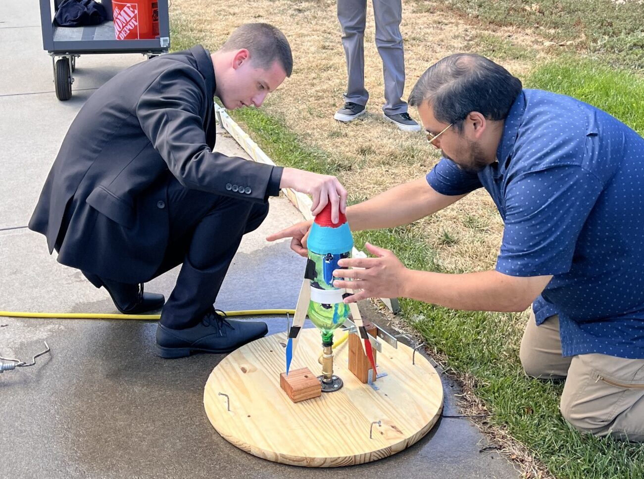 Two men assembling a wooden circular platform with colored mechanical components during an outdoor demonstration or workshop event.