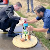 Two men assembling a wooden circular platform with colored mechanical components during an outdoor demonstration or workshop event.