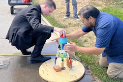 Two men assembling a wooden circular platform with colored mechanical components during an outdoor demonstration or workshop event.