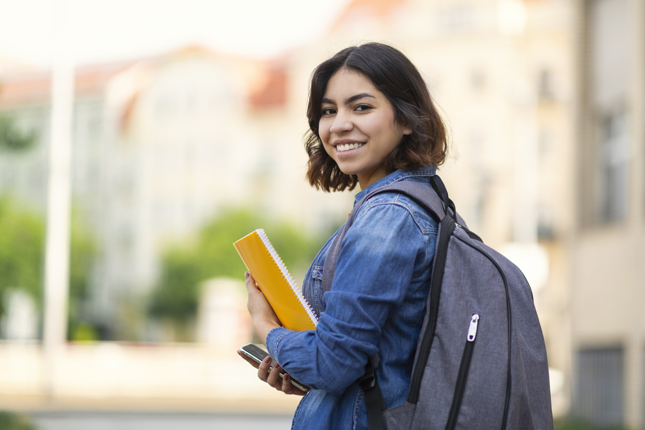 Smiling student with backpack holding yellow notebook and phone outdoors