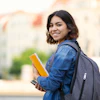 Smiling student with backpack holding yellow notebook and phone outdoors