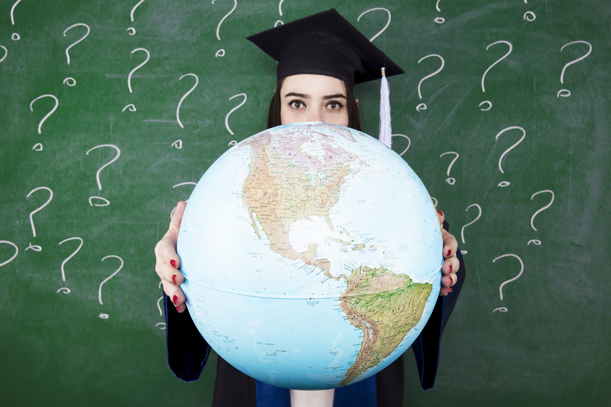 Graduate student in cap and gown holding a large globe in front of a green chalkboard with question marks