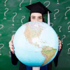 Graduate student in cap and gown holding a large globe in front of a green chalkboard with question marks
