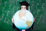 Graduate student in cap and gown holding a large globe in front of a green chalkboard with question marks
