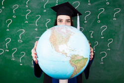 Graduate student in cap and gown holding a large globe in front of a green chalkboard with question marks