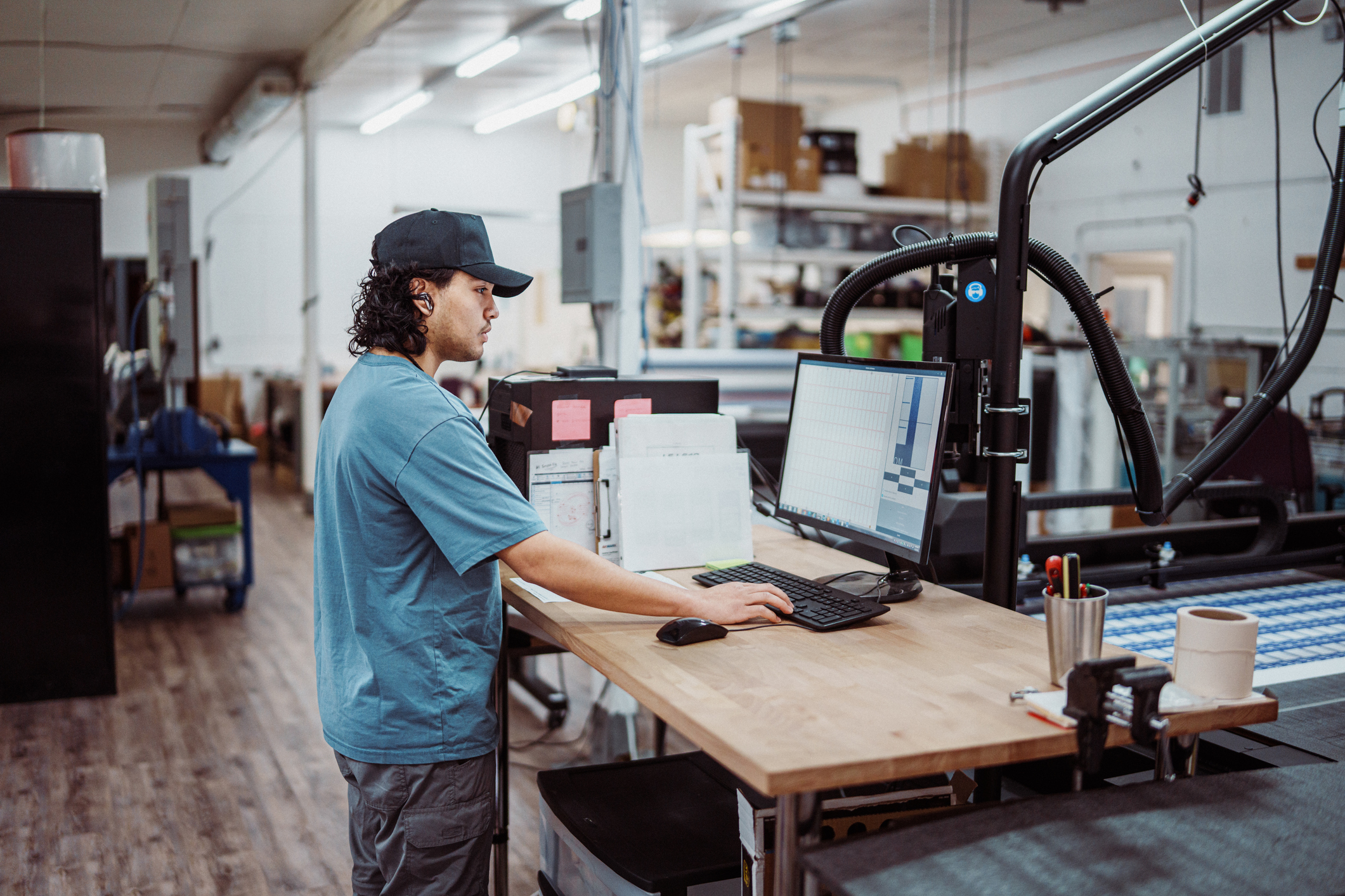 Worker in blue shirt using laptop at wooden desk in maker space with 3D printer and workshop equipment