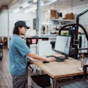 Worker in blue shirt using laptop at wooden desk in maker space with 3D printer and workshop equipment