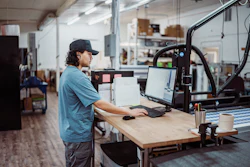 Worker in blue shirt using laptop at wooden desk in maker space with 3D printer and workshop equipment