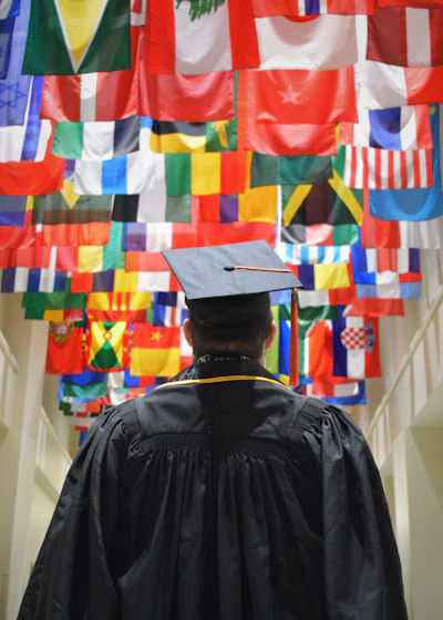 Graduate in black academic regalia standing before a wall of colorful international flags representing global education and diversity