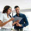 Two professionals smiling while reviewing content on a tablet in a modern office with city views