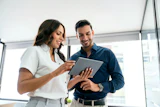 Two professionals smiling while reviewing content on a tablet in a modern office with city views