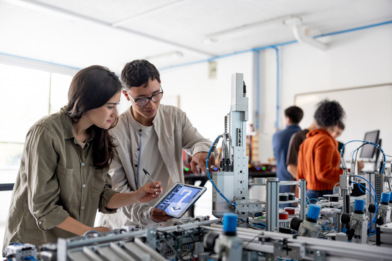 Two engineers collaborating at a robotic workstation in a laboratory, reviewing data on tablet while examining mechanical equipment.