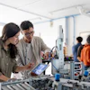 Two engineers collaborating at a robotic workstation in a laboratory, reviewing data on tablet while examining mechanical equipment.