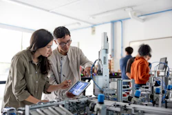 Two engineers collaborating at a robotic workstation in a laboratory, reviewing data on tablet while examining mechanical equipment.