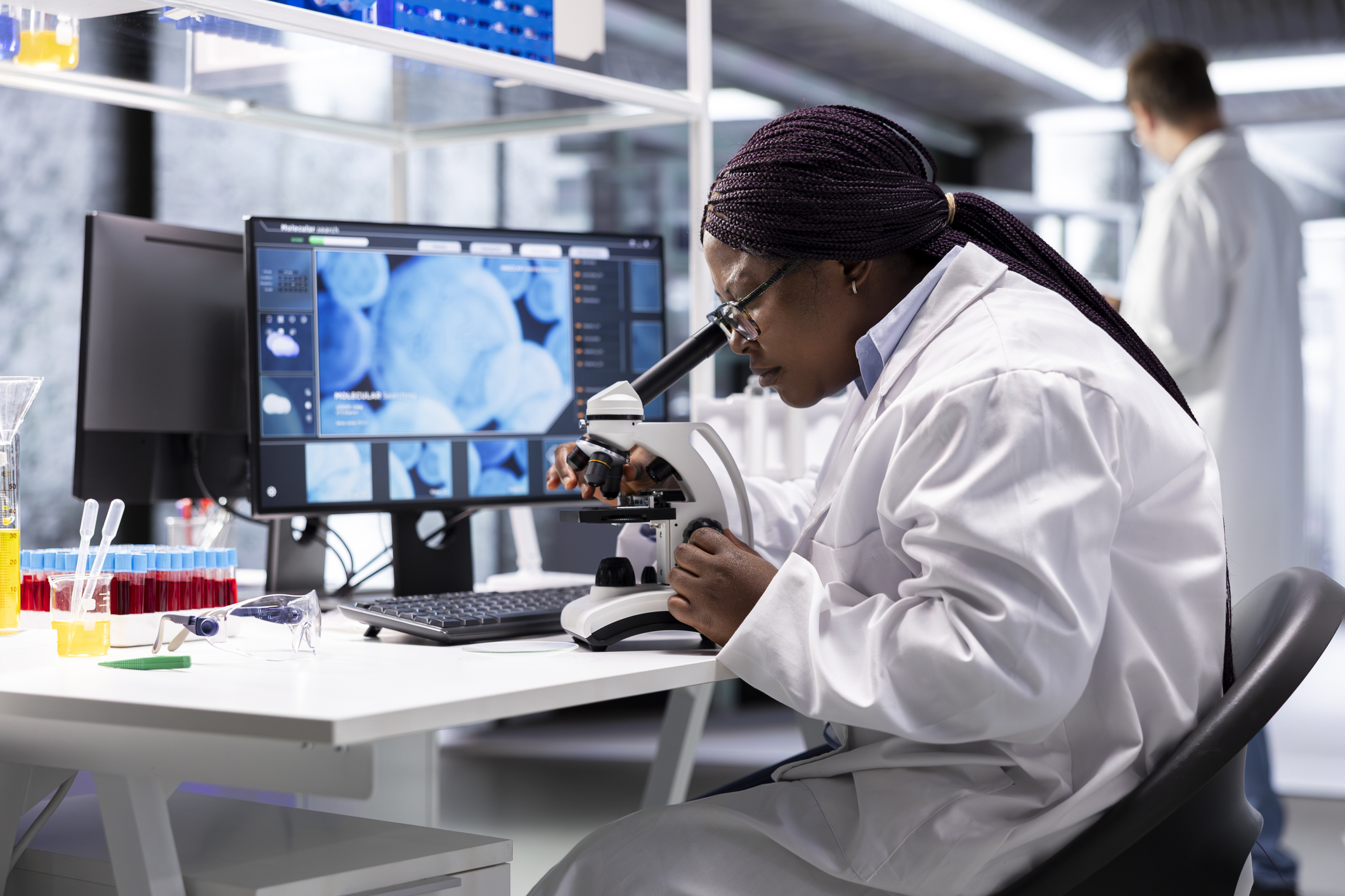 Female scientist examining samples under microscope in modern laboratory with medical imaging displayed on monitor