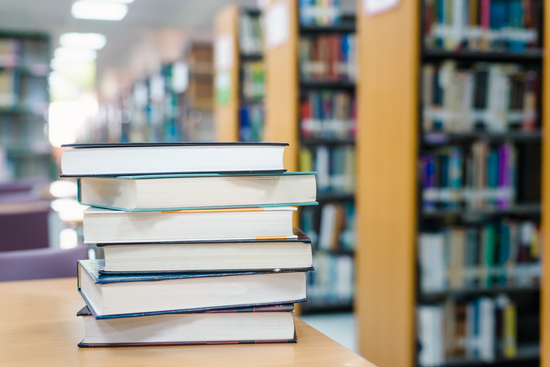 Stack of books on a wooden table in a library with shelves blurred in the background