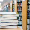 Stack of books on a wooden table in a library with shelves blurred in the background