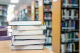 Stack of books on a wooden table in a library with shelves blurred in the background