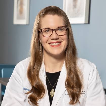 Professional headshot of a smiling woman wearing glasses and a white medical coat with credentials