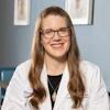 Professional headshot of a smiling woman wearing glasses and a white medical coat with credentials