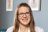 Professional headshot of a smiling woman wearing glasses and a white medical coat with credentials