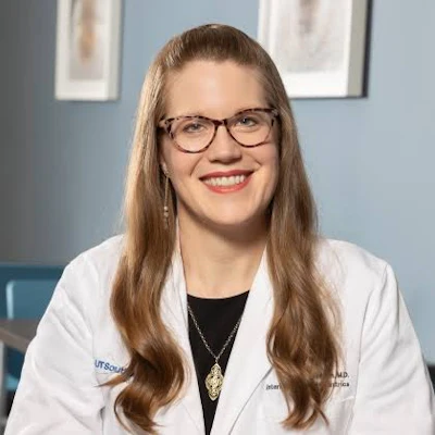 Professional headshot of a smiling woman wearing glasses and a white medical coat with credentials