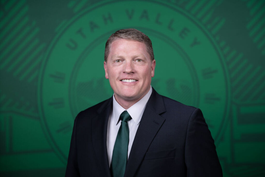 Professional headshot of a smiling man in dark blazer and green tie against green institutional background