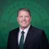 Professional headshot of a smiling man in dark blazer and green tie against green institutional background