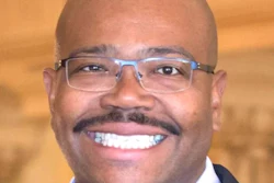 Professional headshot of a smiling man wearing blue-framed glasses and dark business suit