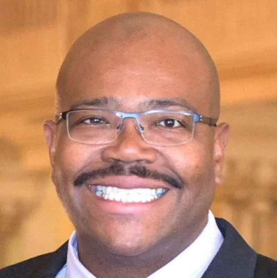 Professional headshot of a smiling man wearing blue-framed glasses and dark business suit