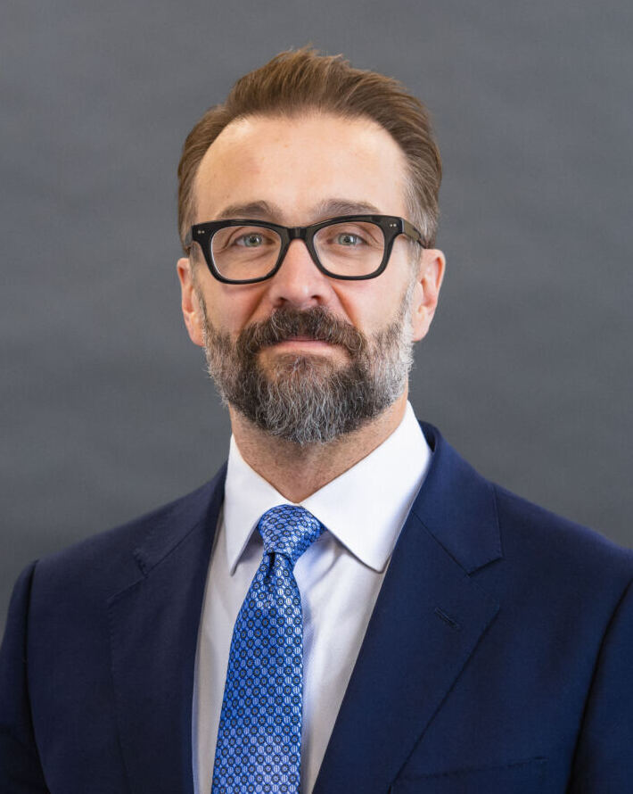Professional headshot of bearded man wearing glasses, navy blazer, and blue tie against gray background