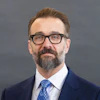Professional headshot of bearded man wearing glasses, navy blazer, and blue tie against gray background