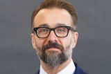 Professional headshot of bearded man wearing glasses, navy blazer, and blue tie against gray background