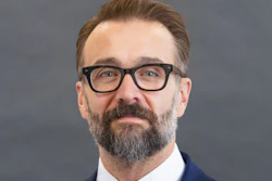 Professional headshot of bearded man wearing glasses, navy blazer, and blue tie against gray background