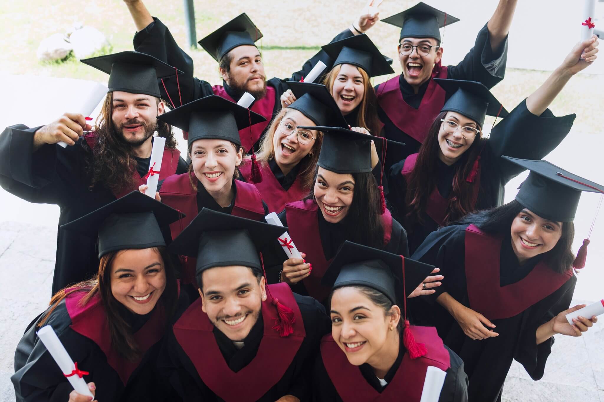Group of happy graduates in burgundy robes and mortarboards celebrating together with raised arms and diplomas
