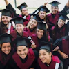 Group of happy graduates in burgundy robes and mortarboards celebrating together with raised arms and diplomas