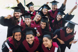 Group of happy graduates in burgundy robes and mortarboards celebrating together with raised arms and diplomas