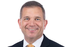 Professional headshot of a smiling man in business attire with dark suit and gold tie