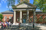 Historic brick building with classical columns and portico, surrounded by visitors on a sunny day