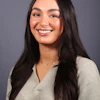 Professional headshot of a smiling woman with long dark hair wearing a beige ribbed sweater and delicate necklace against a gray background