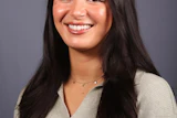 Professional headshot of a smiling woman with long dark hair wearing a beige ribbed sweater and delicate necklace against a gray background