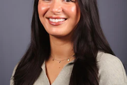 Professional headshot of a smiling woman with long dark hair wearing a beige ribbed sweater and delicate necklace against a gray background