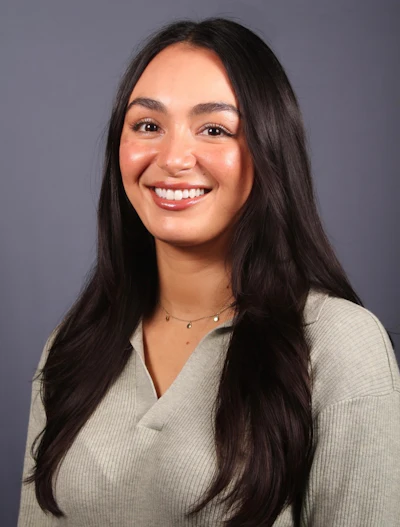 Professional headshot of a smiling woman with long dark hair wearing a beige ribbed sweater and delicate necklace against a gray background