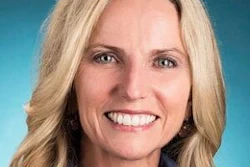 Professional headshot of a smiling woman with blonde wavy hair wearing a light blue shirt against a teal background