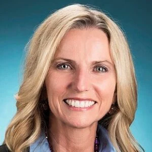 Professional headshot of a smiling woman with blonde wavy hair wearing a light blue shirt against a teal background