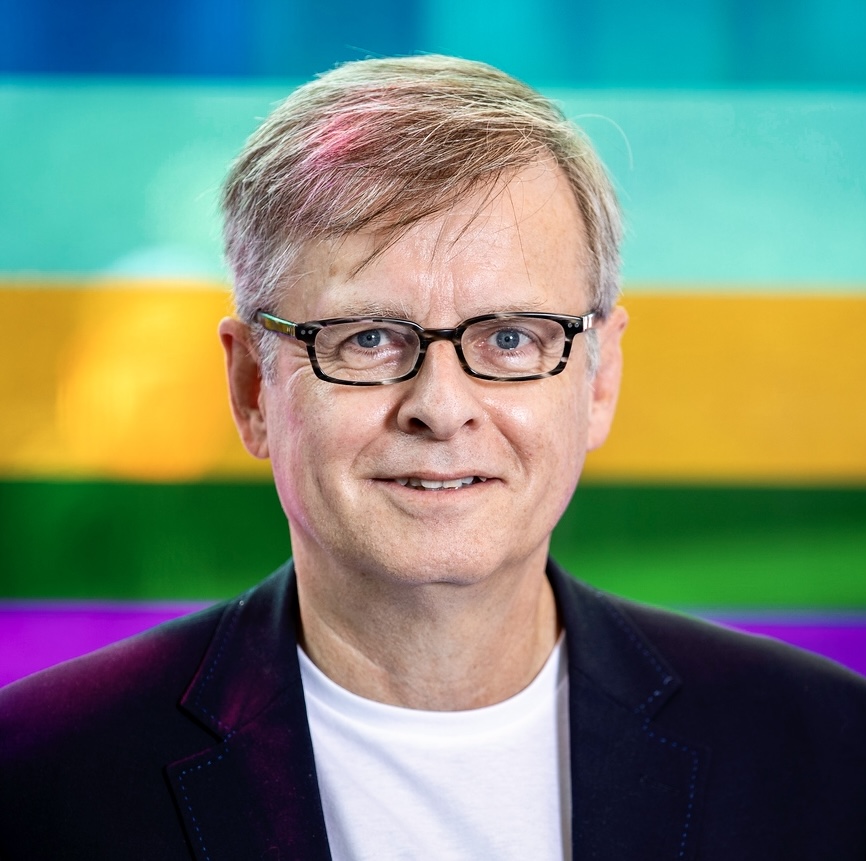 Professional headshot of a man with glasses smiling against a vibrant rainbow gradient background Headshot