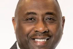 Professional headshot of a smiling man in a dark suit and white shirt against a light background
